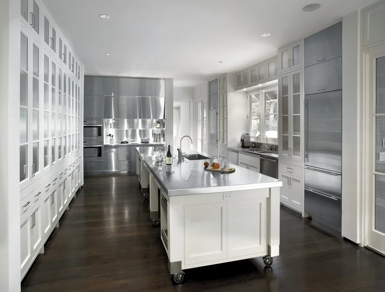 The Riverbank Residence kitchen features a long central island with a stainless steel countertop and tall cabinetry with glass doors.