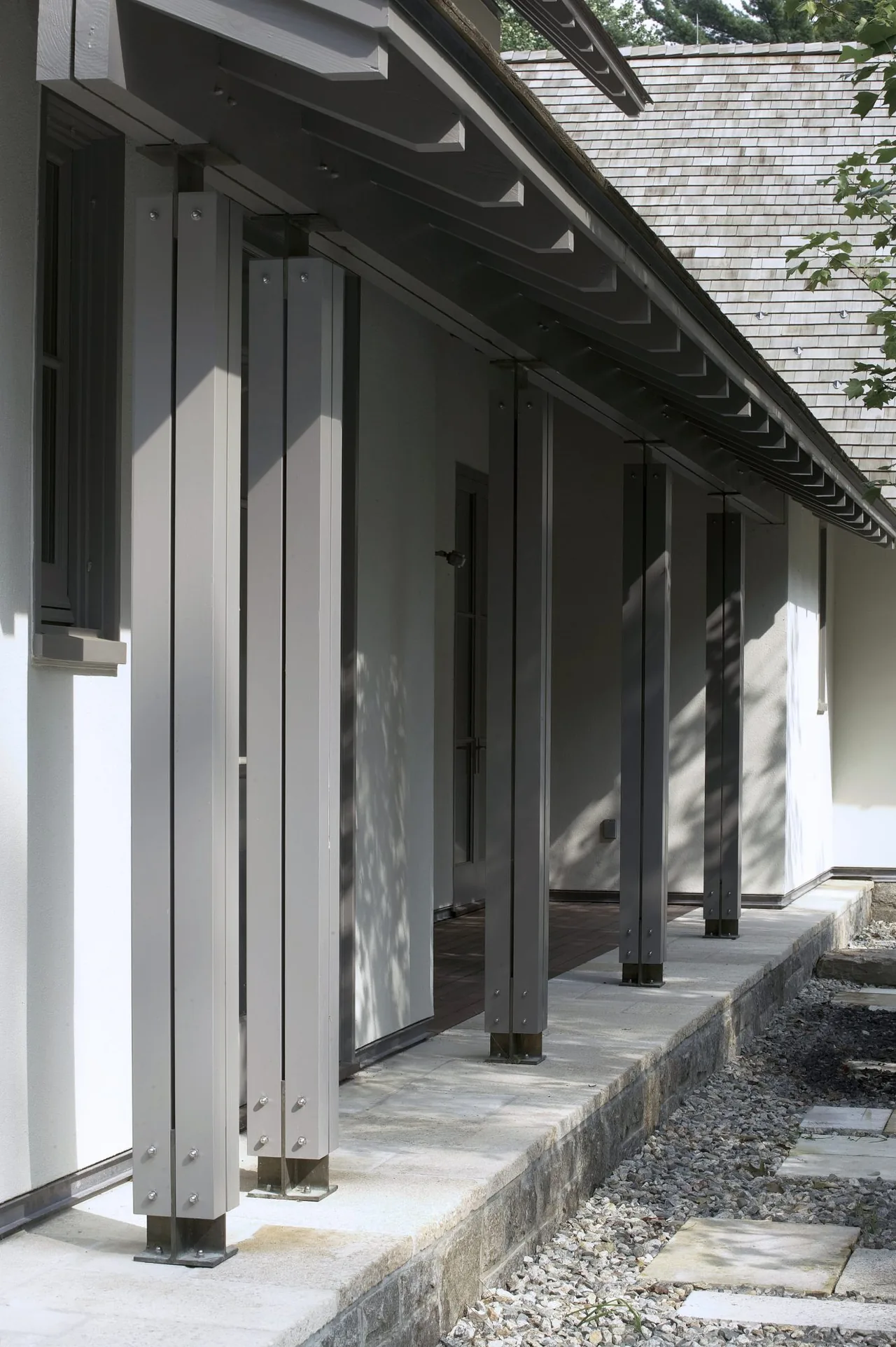 Riverbank Residence porch features vertical steel columns on a stone retaining wall base with a gravel path.