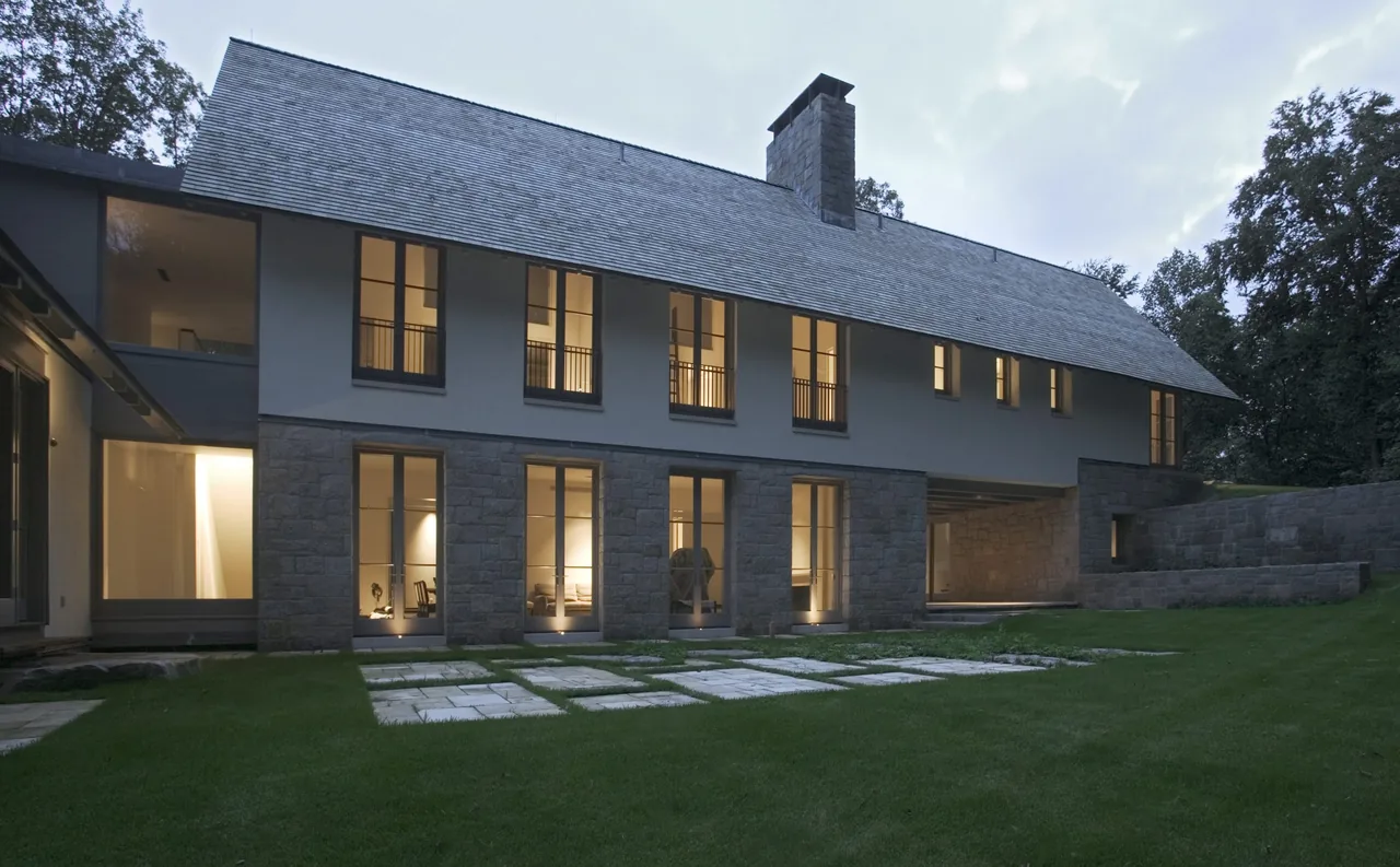 Riverbank Residence features stone lower walls, multiple vertical windows, and a steep roof pitch with a chimney.