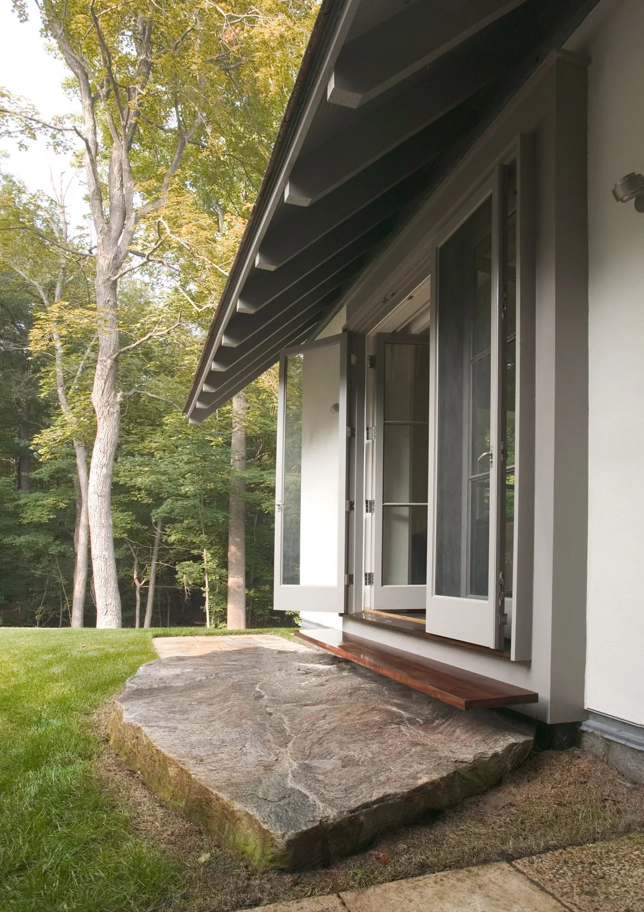 Exterior view of open glass double doors leading to a natural stone patio and wooden bench at Riverbank Residence.