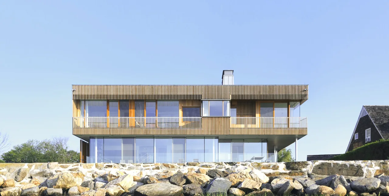 Spiral House features a two-story design with vertical wood slat siding and floor-to-ceiling glass on the lower level, elevated on a rocky foundation.