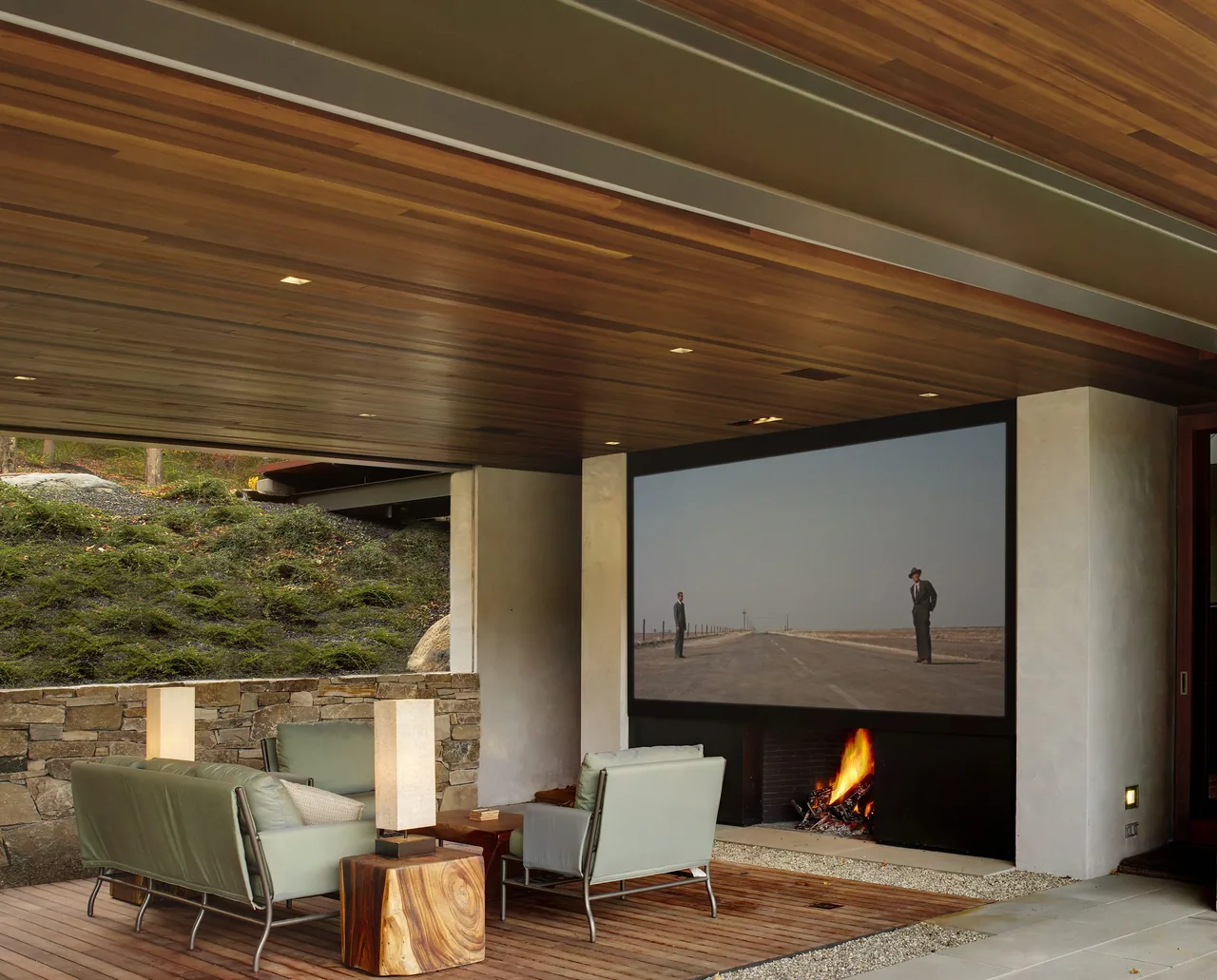 Covered outdoor living space features built-in fireplace, wooden ceiling, stone wall, and a large projection screen for entertainment.