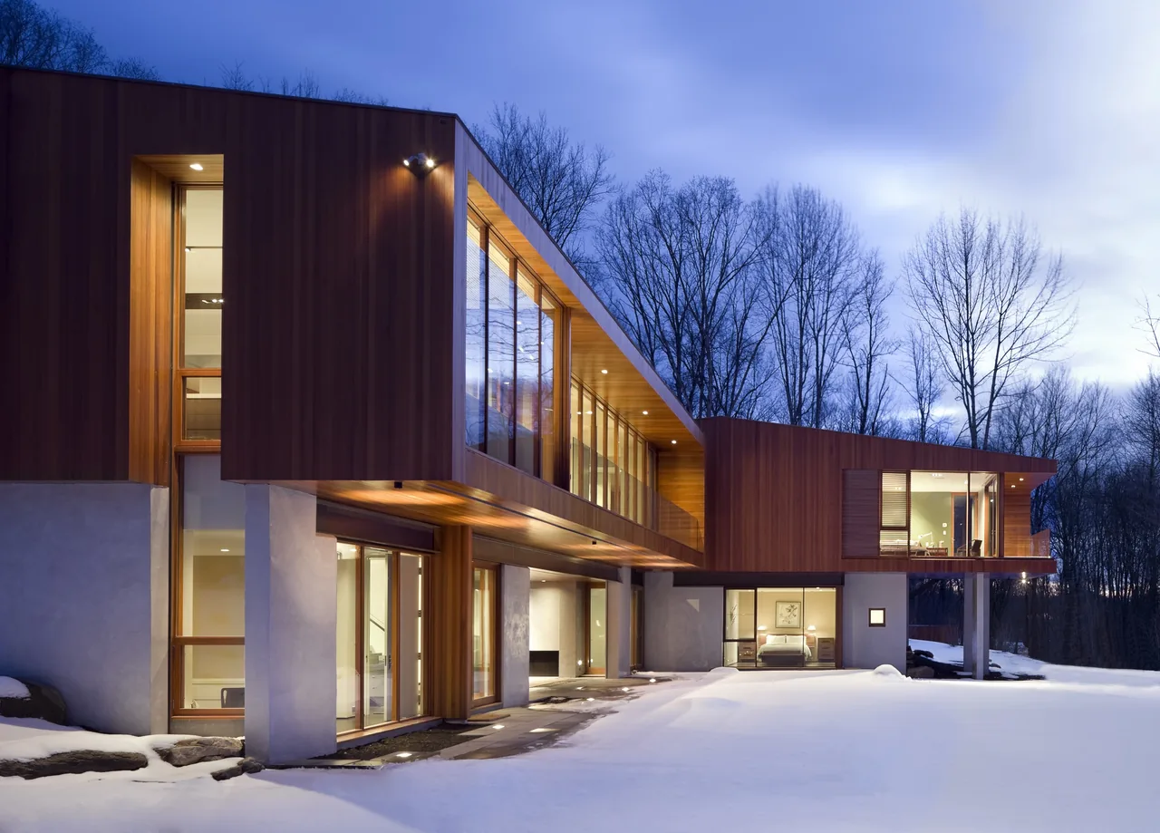Bridge House features wood siding and large glass windows in a snowy landscape with surrounding leafless trees.