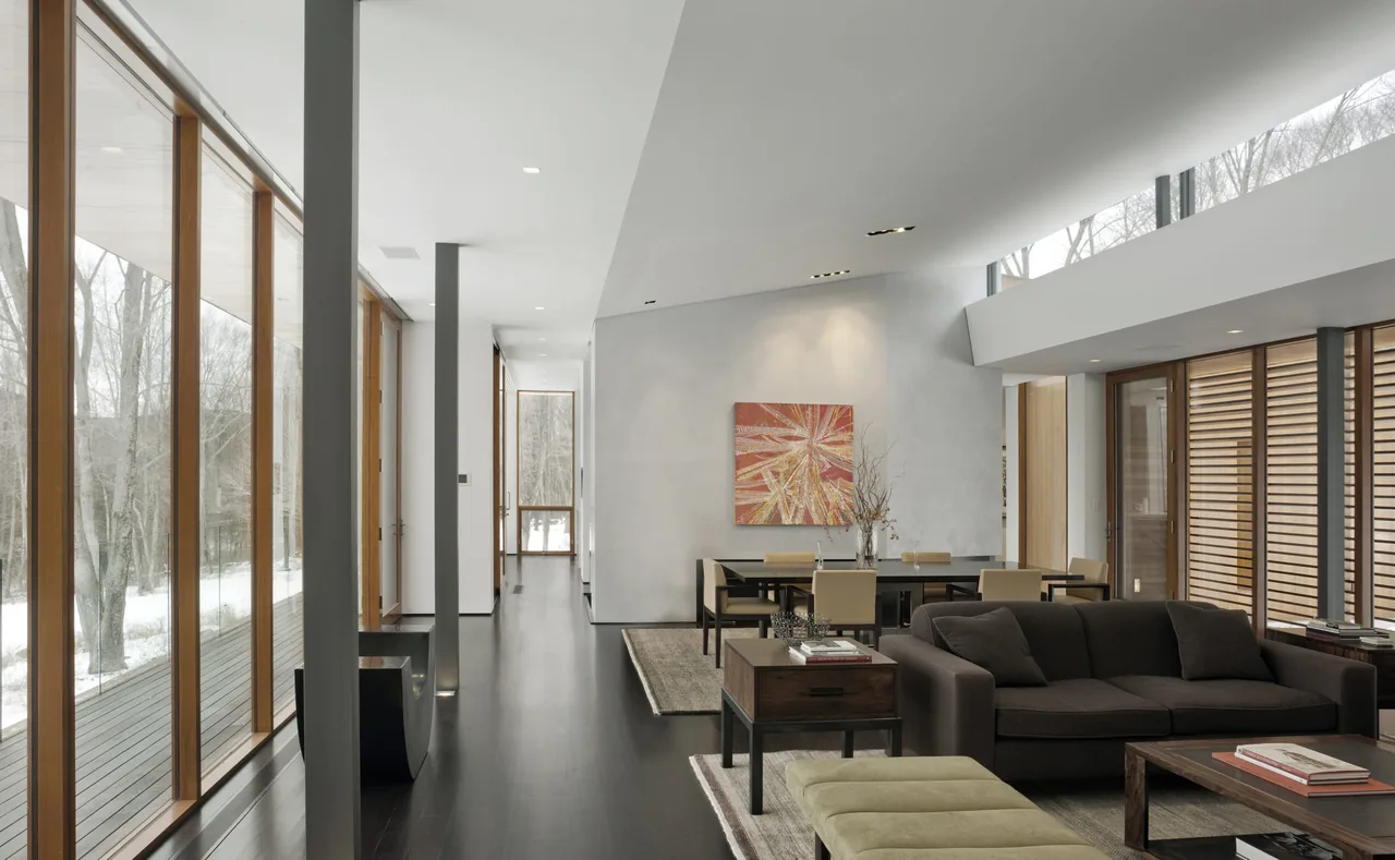 Open plan living and dining area with dark wood flooring, wood window frames, and large windows showing a winter landscape.