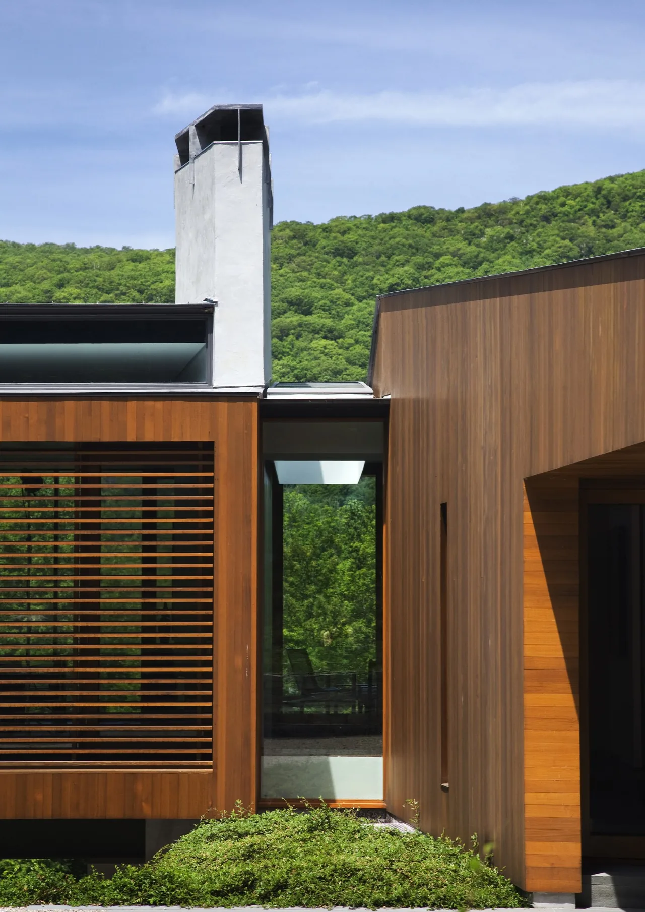 Bridge House features horizontal wood siding and a wood slat screen next to a white chimney and a glass window reflecting greenery.