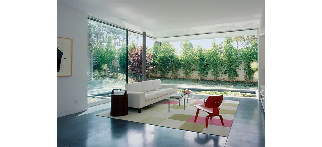 Ehrlich House living room with large glass windows overlooking a garden and lily pond, featuring a colorful area rug and minimalist furniture.