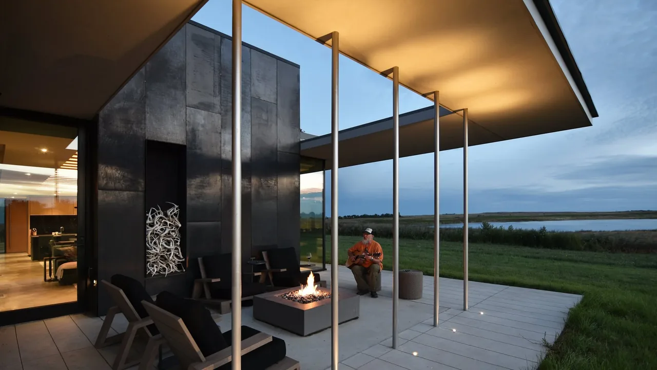 Patio at Spoonbill Ranch features a fire pit, tall metal wall, and a covered seating area overlooking a pond in the evening light.