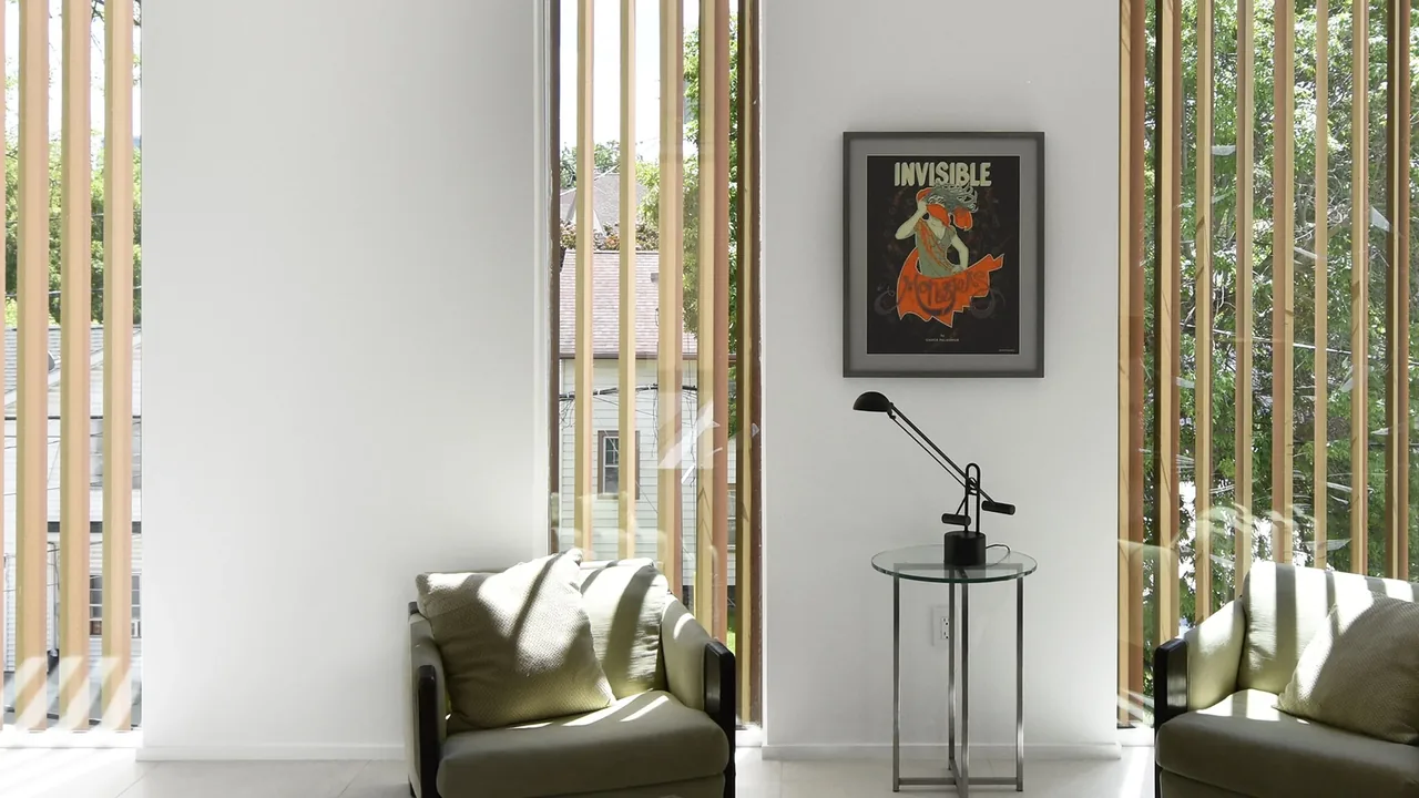 Living room corner with two chairs, a glass side table with a black desk lamp, vertical wooden slats on windows, and framed artwork.