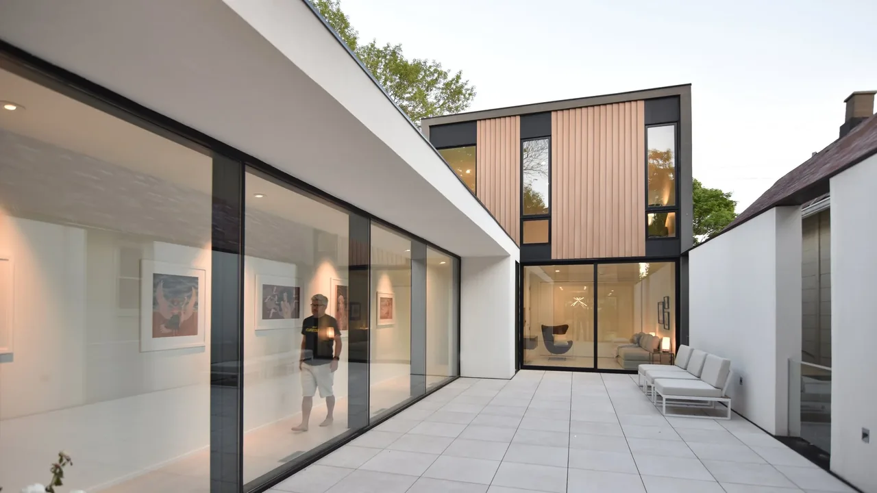 Exterior rear view of Curtain House with vertical wood siding, expansive glazing, and a tiled courtyard patio with seating.