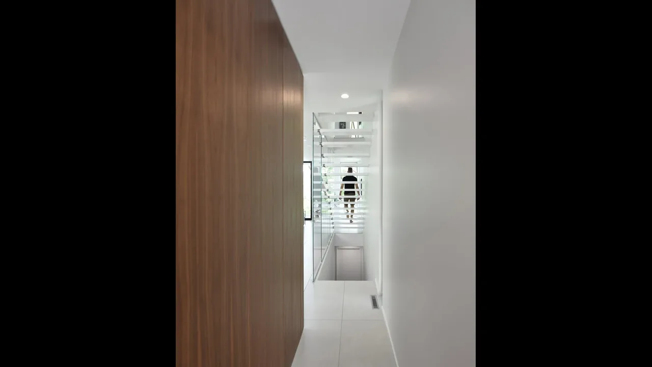 A narrow hallway with a combination of wood and white walls, featuring glass railings and horizontal slats near a stairway.