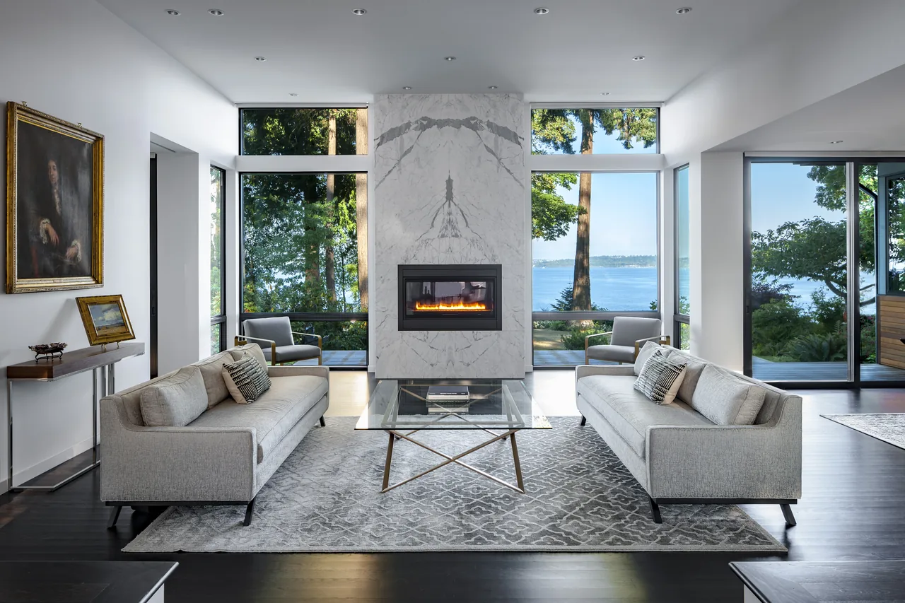 The Sunrise House living room features floor-to-ceiling windows framing water views and a marble fireplace centerpiece.