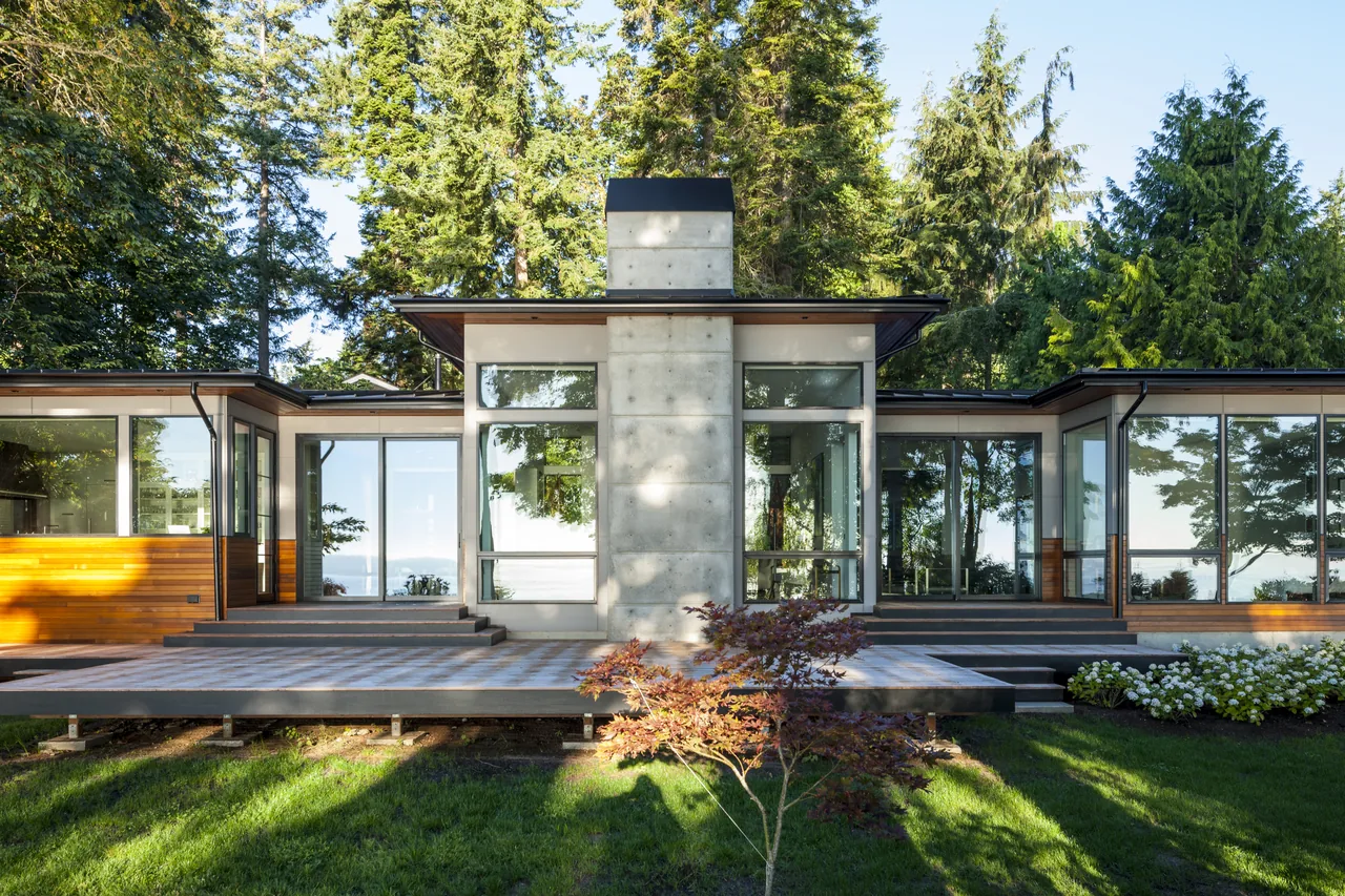 Exterior rear view of Sunrise House showing concrete chimney, extensive glazing, and wood siding with forest backdrop.