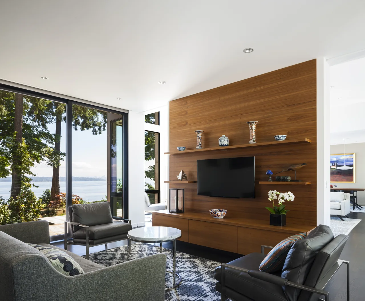 Living room in Sunrise House features a wood-paneled wall with TV, large windows facing water, and modern seating.