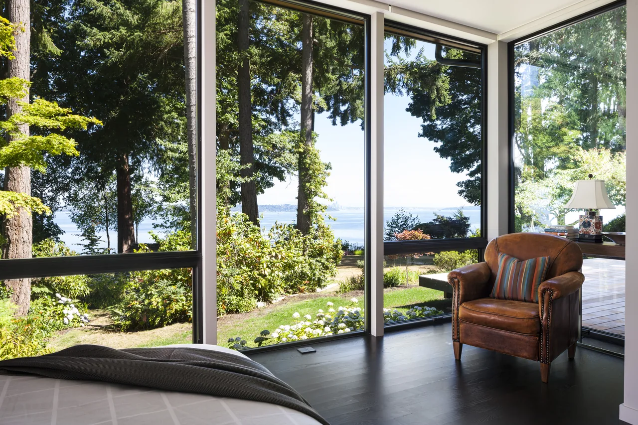 Bedroom corner with large floor-to-ceiling windows providing views of trees and water.