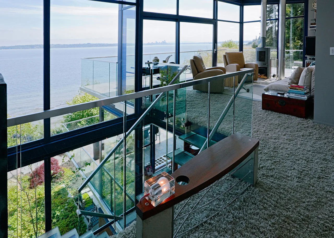 Living area with large glass windows and a glass-railing staircase overlooking a water view.