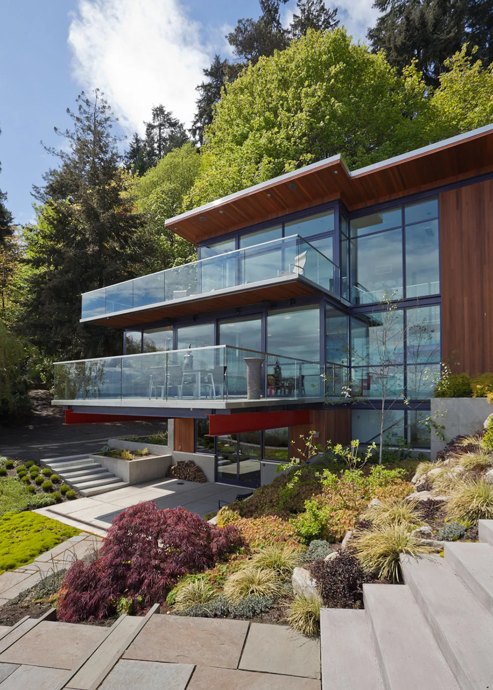 The Bainbridge Island Guest Residence features floor-to-ceiling windows, wood siding, and glass guardrails on cantilevered balconies overlooking a landscaped yard.