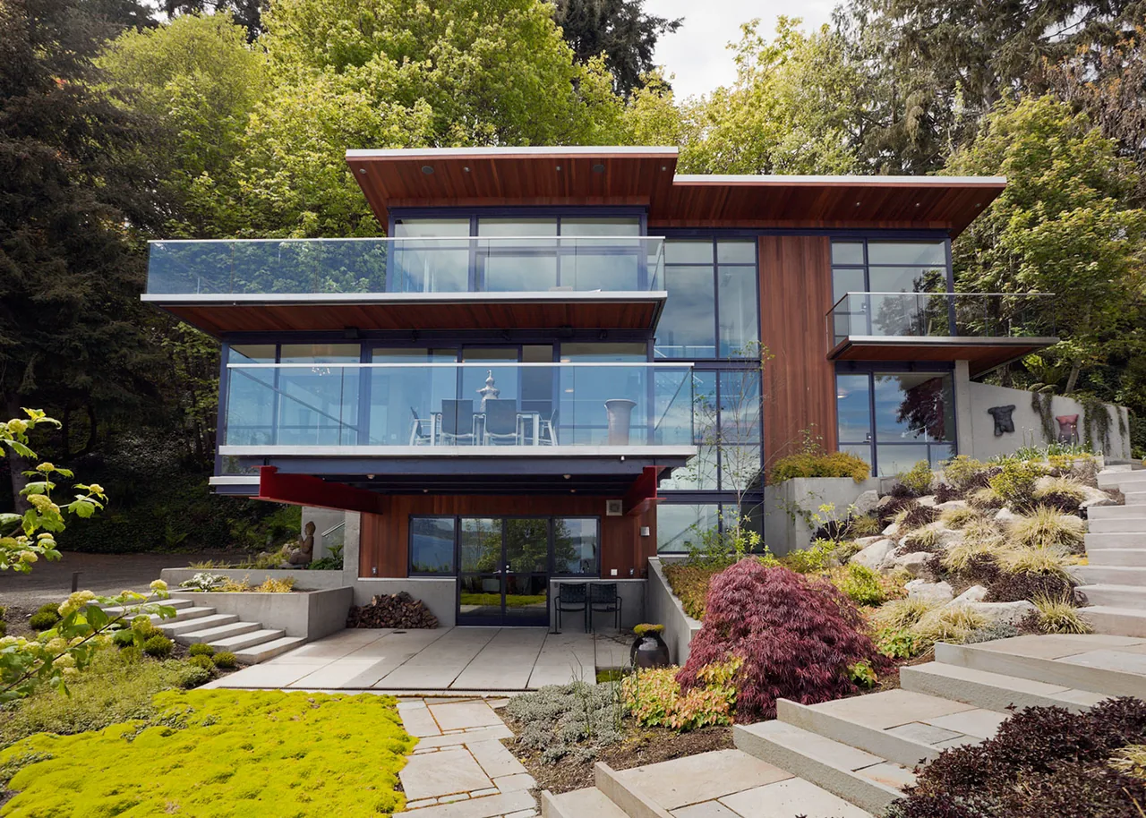Bainbridge Island Guest Residence features glass balcony railings and wood siding, situated on a landscaped hillside.