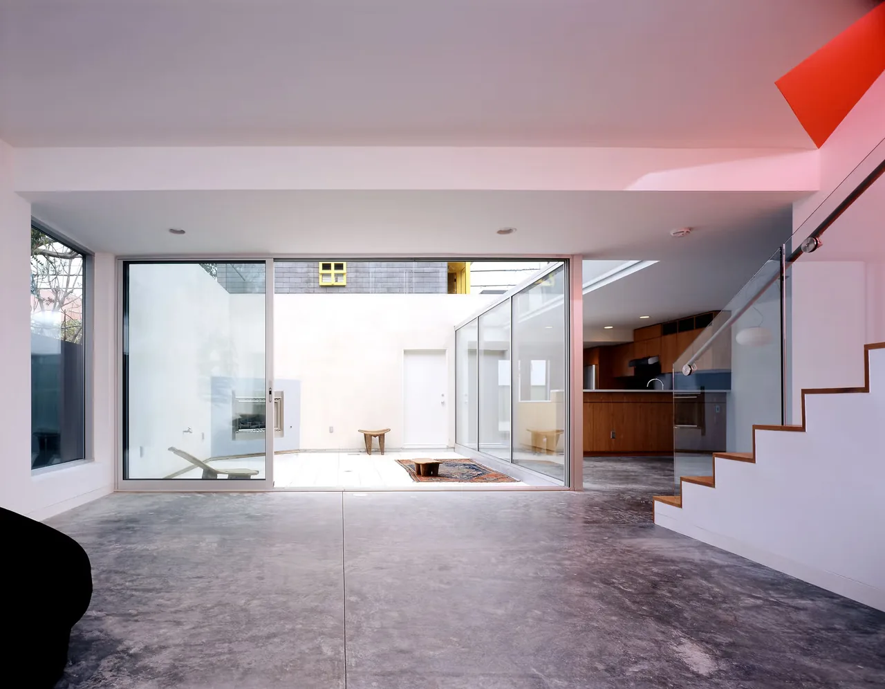 Modern interior with concrete floor, glass sliding doors leading to a small courtyard, and wooden kitchen cabinetry visible in background.