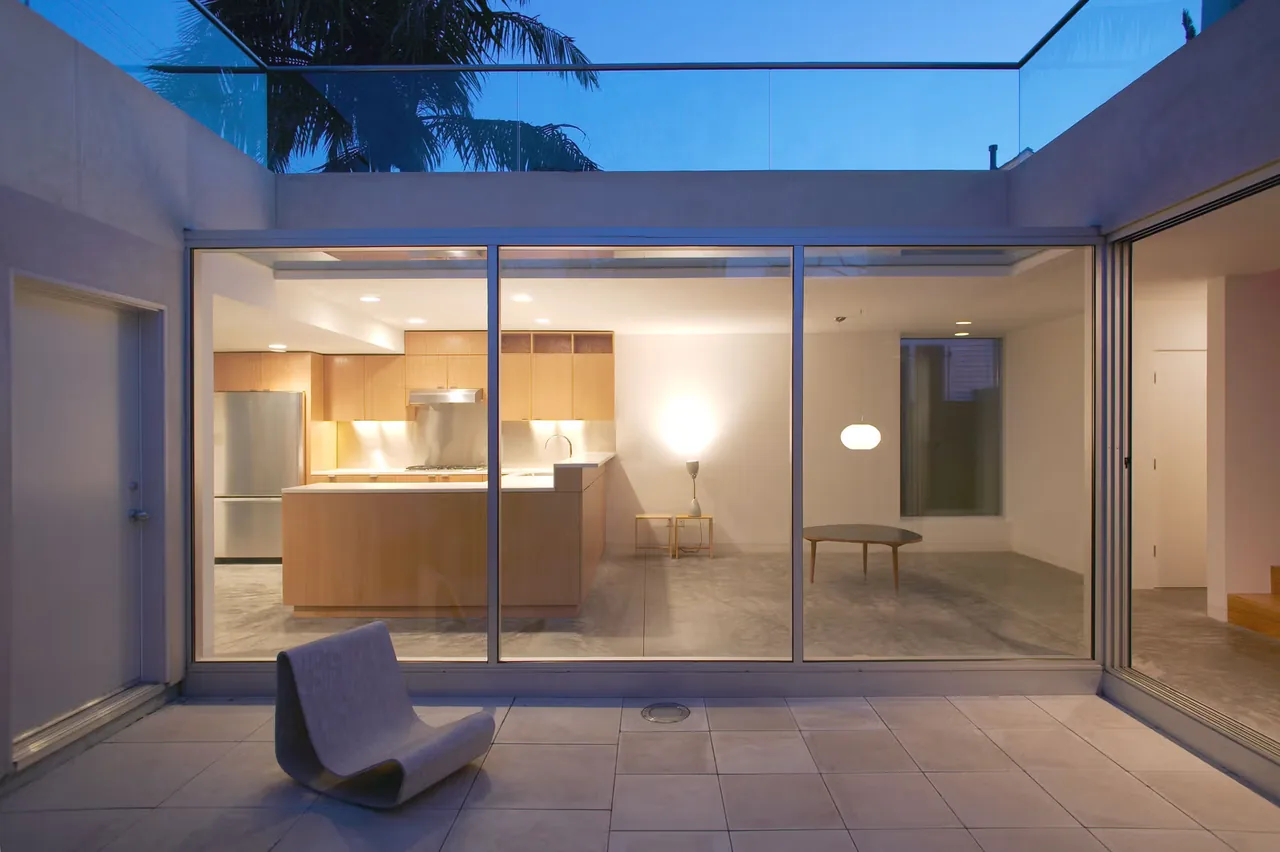 Evening view of a modern kitchen and living room through floor-to-ceiling glass doors opening to a courtyard patio.