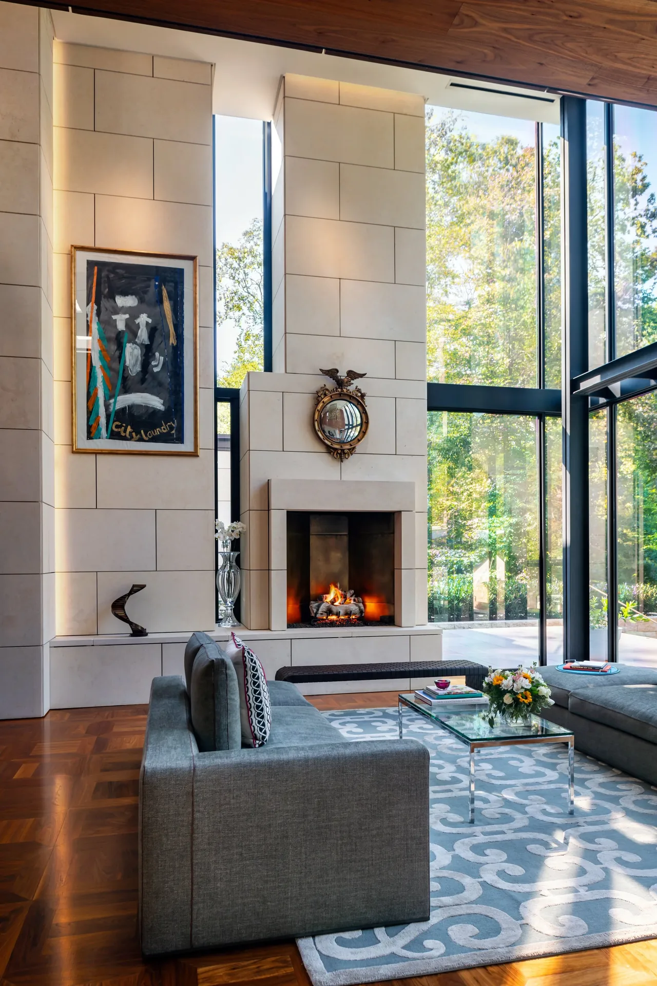 Modern living room features a large stone fireplace and expansive floor-to-ceiling windows providing a view of outdoor trees.