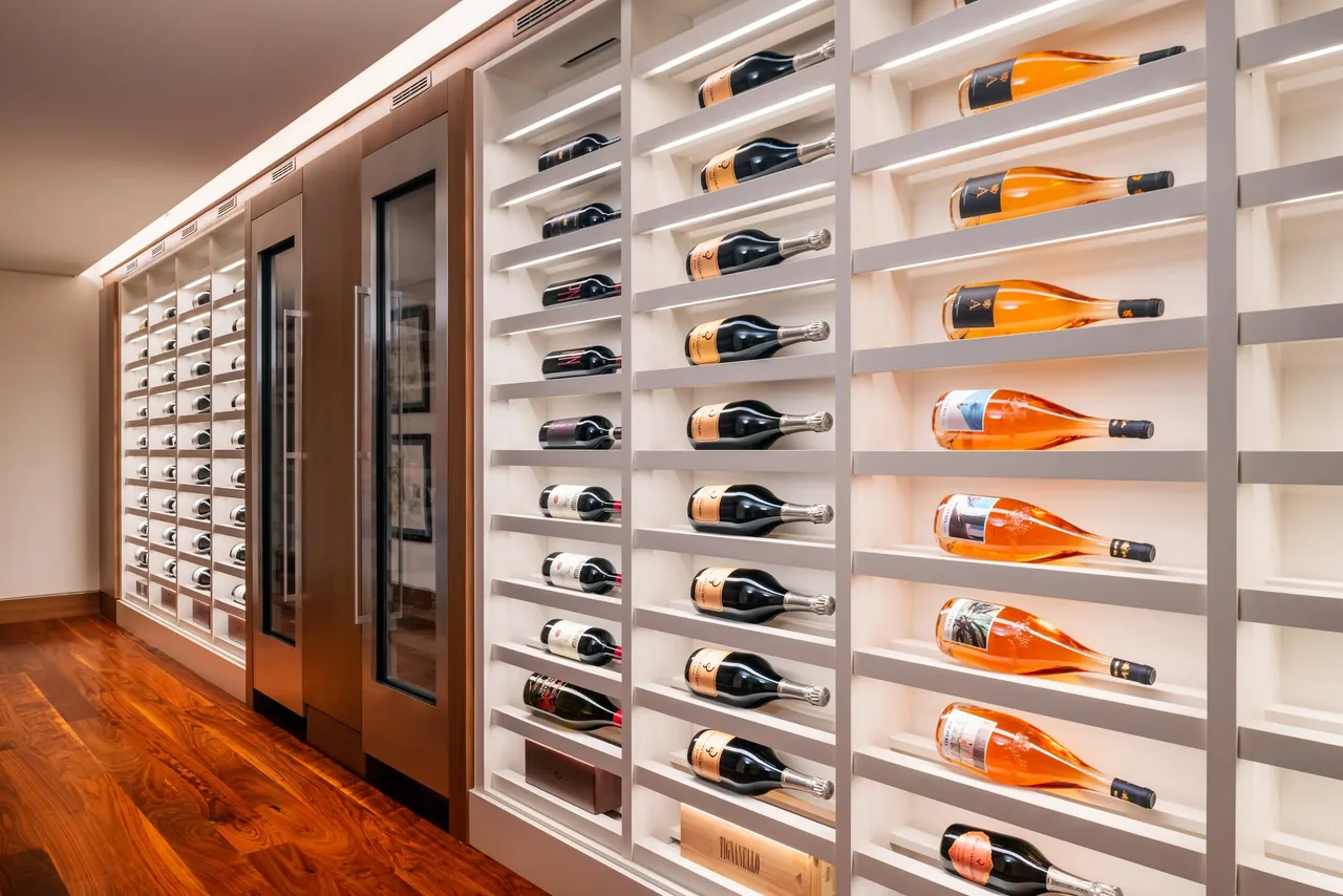 Wine bottles stored horizontally on built-in racks alongside two stainless steel wine refrigerators on a hardwood floor.