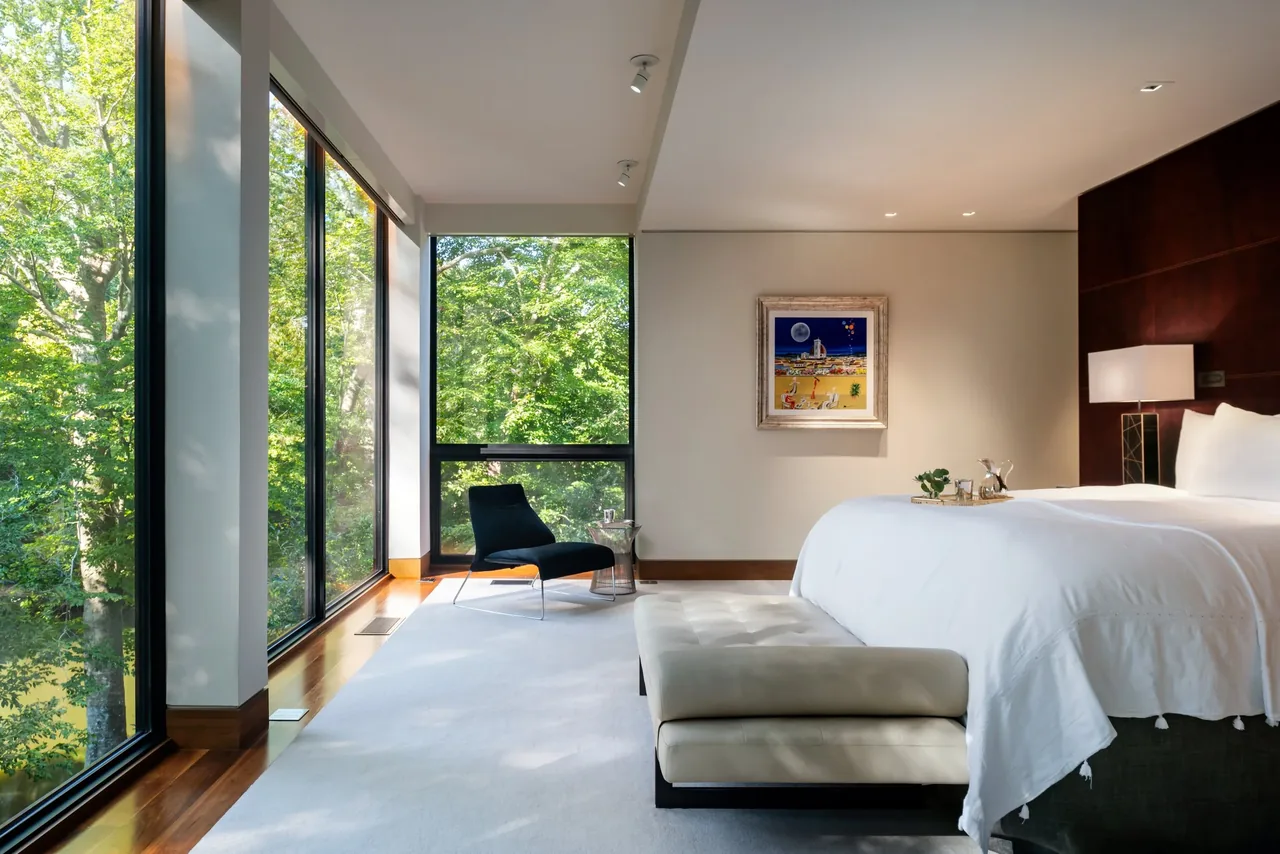 Bedroom features floor-to-ceiling glazing, a black lounge chair, and white bedding with a leather bench at the foot.