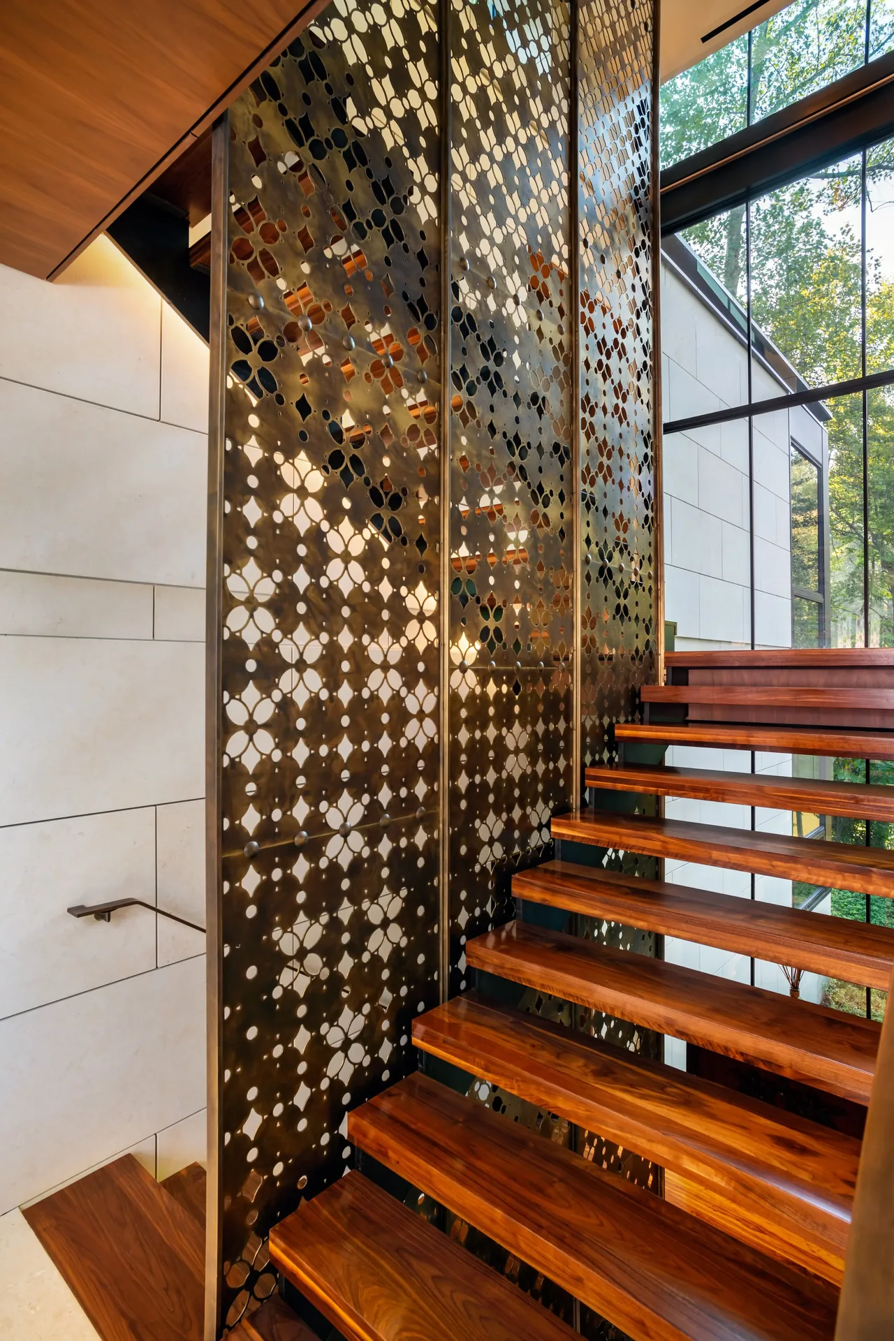 Floating wooden stairs with a decorative perforated metal panel and floor-to-ceiling windows in this modern interior.