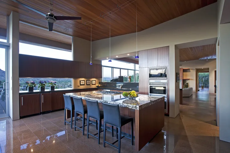 Monks Shadow Residence kitchen features a granite island, wood cabinetry, and expansive windows for natural light and desert views.