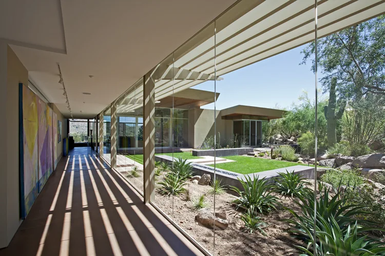 The Monks Shadow Residence features a glass-walled hallway with views of a desert courtyard landscape.