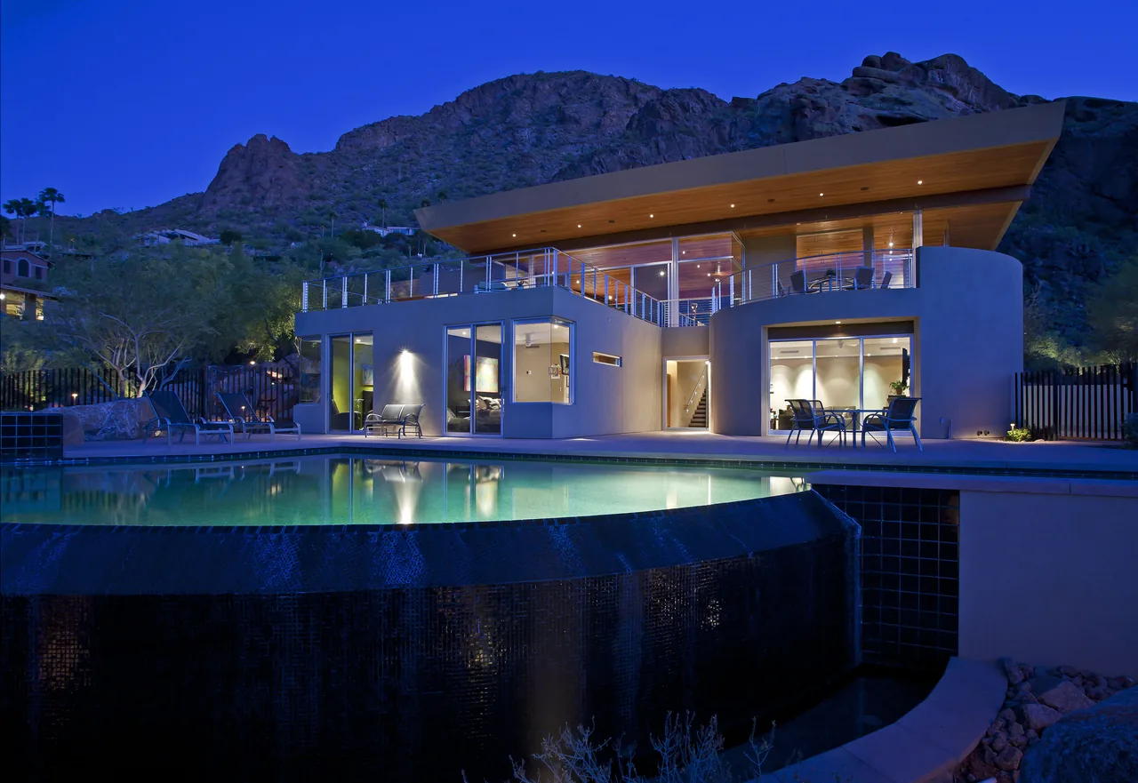 Two-story modern house with a cantilevered wood-paneled roof over outdoor seating and an illuminated pool with waterfall