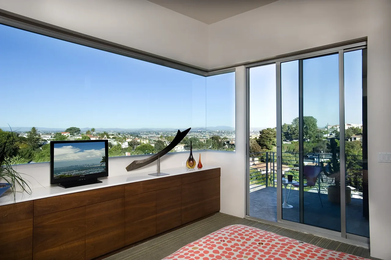Bedroom corner with floor-to-ceiling corner window and sliding glass door opens to an outdoor balcony with city views.
