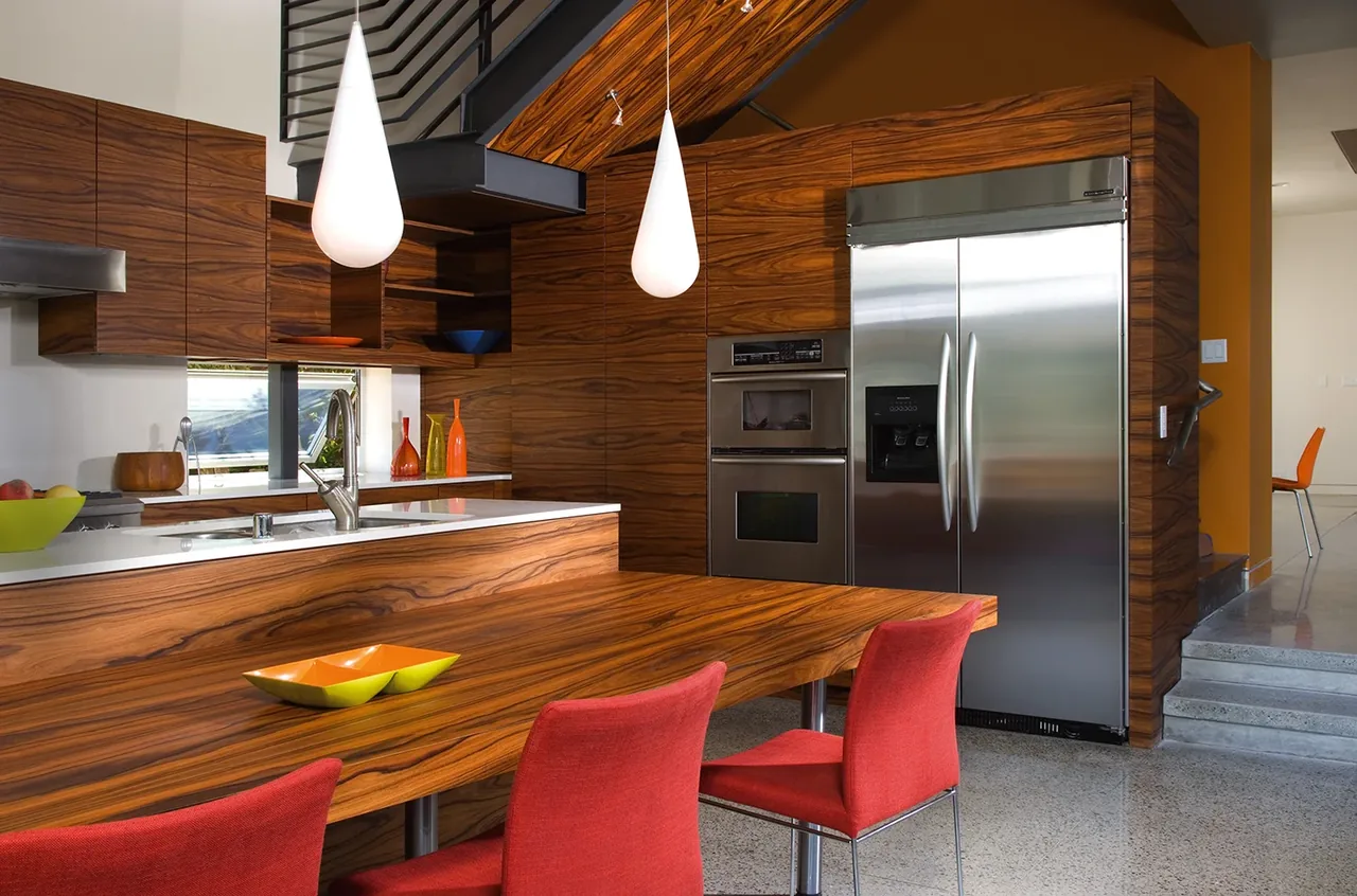 Casa Familia kitchen features rich wood cabinetry, stainless steel fridge and ovens, and a wood table with red chairs.