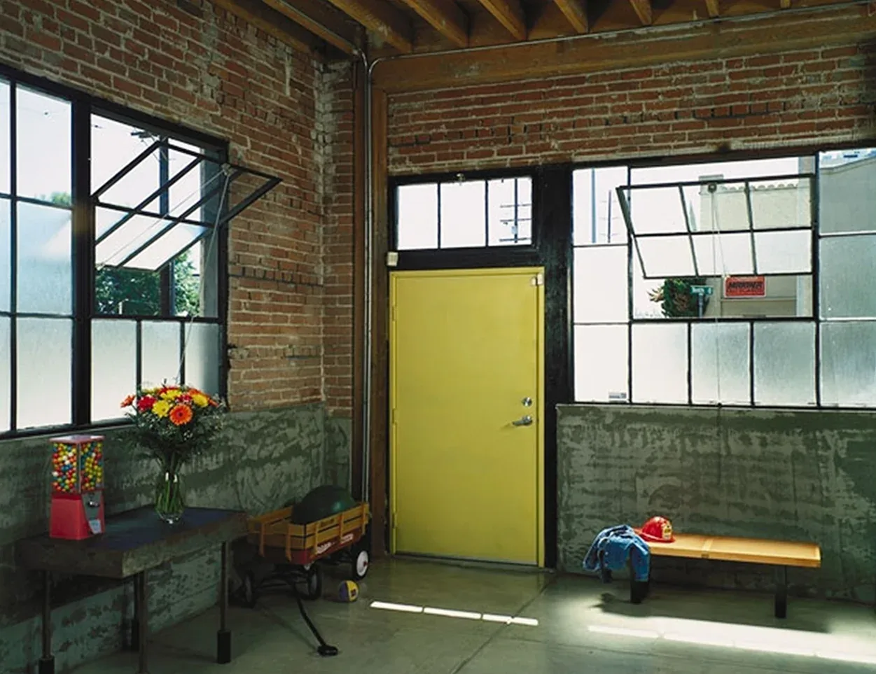 This industrial loft space features exposed brick and concrete walls, a yellow door, and black metal window frames with ventilating panes.