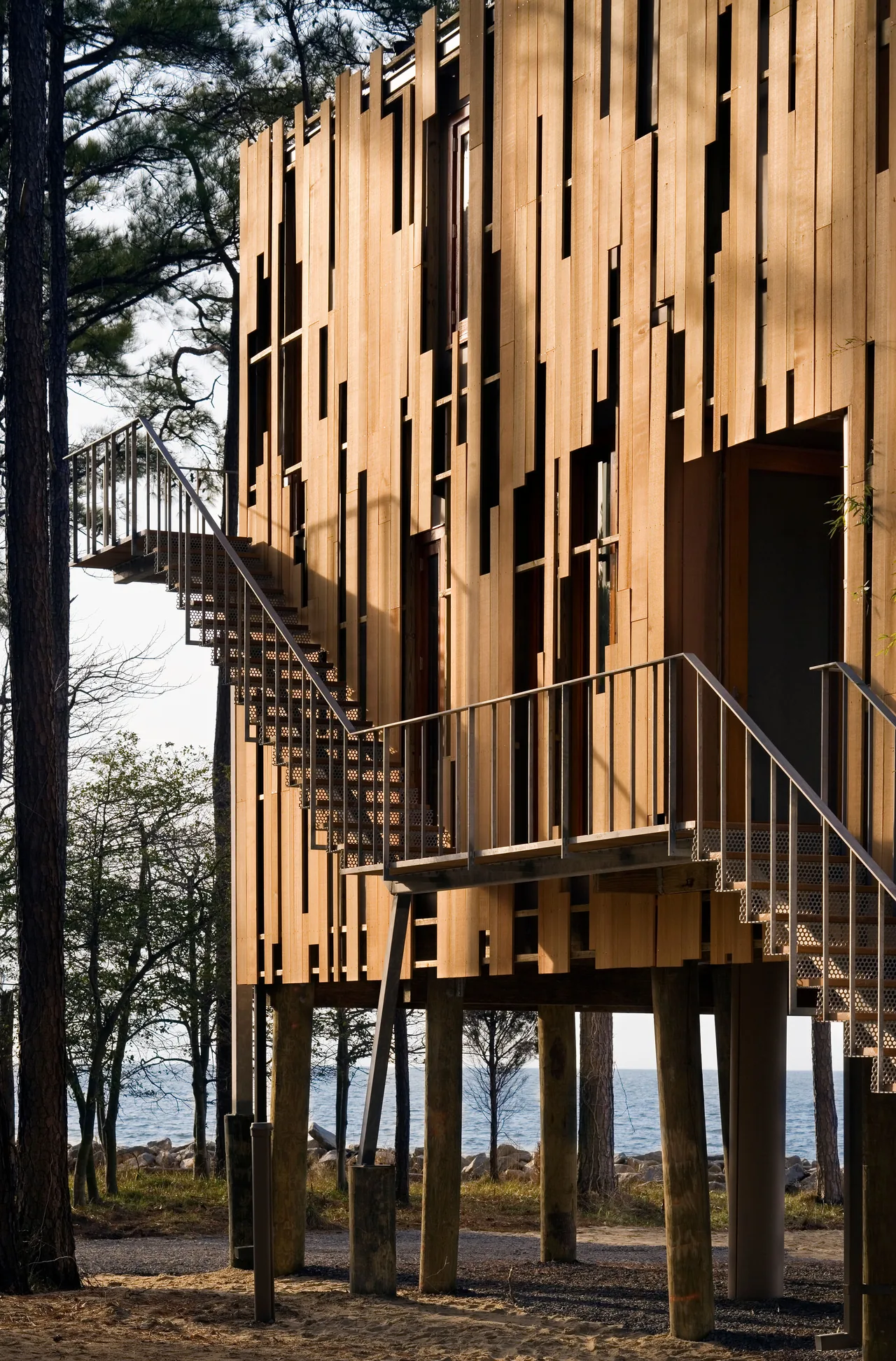 The Loblolly House features vertical wooden siding and an external metal staircase, elevated on wooden pilings by the water.