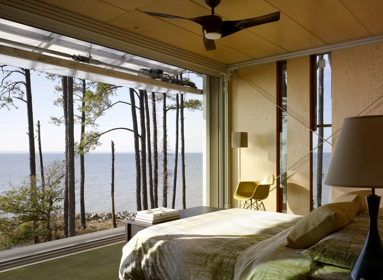 Bedroom of Loblolly House features large window framing a waterfront view with woods beyond.