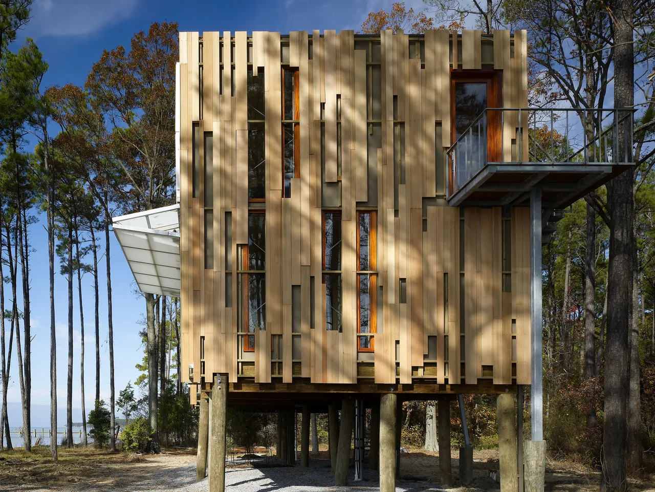 Loblolly House features vertical wood paneling and is elevated on stilts above a gravel ground surface.