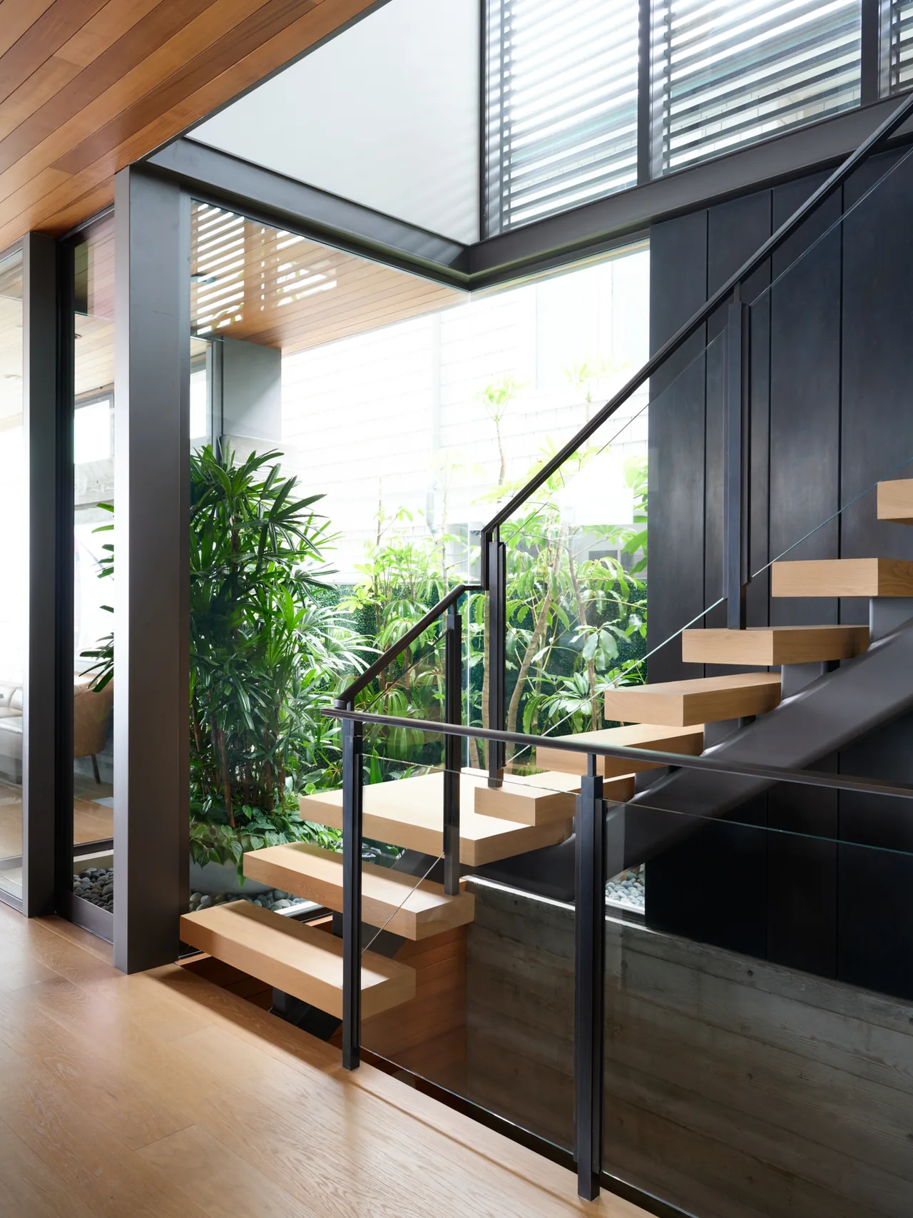 Floating wood staircase with glass railing and black metal supports next to a garden viewed through floor-to-ceiling glazing.