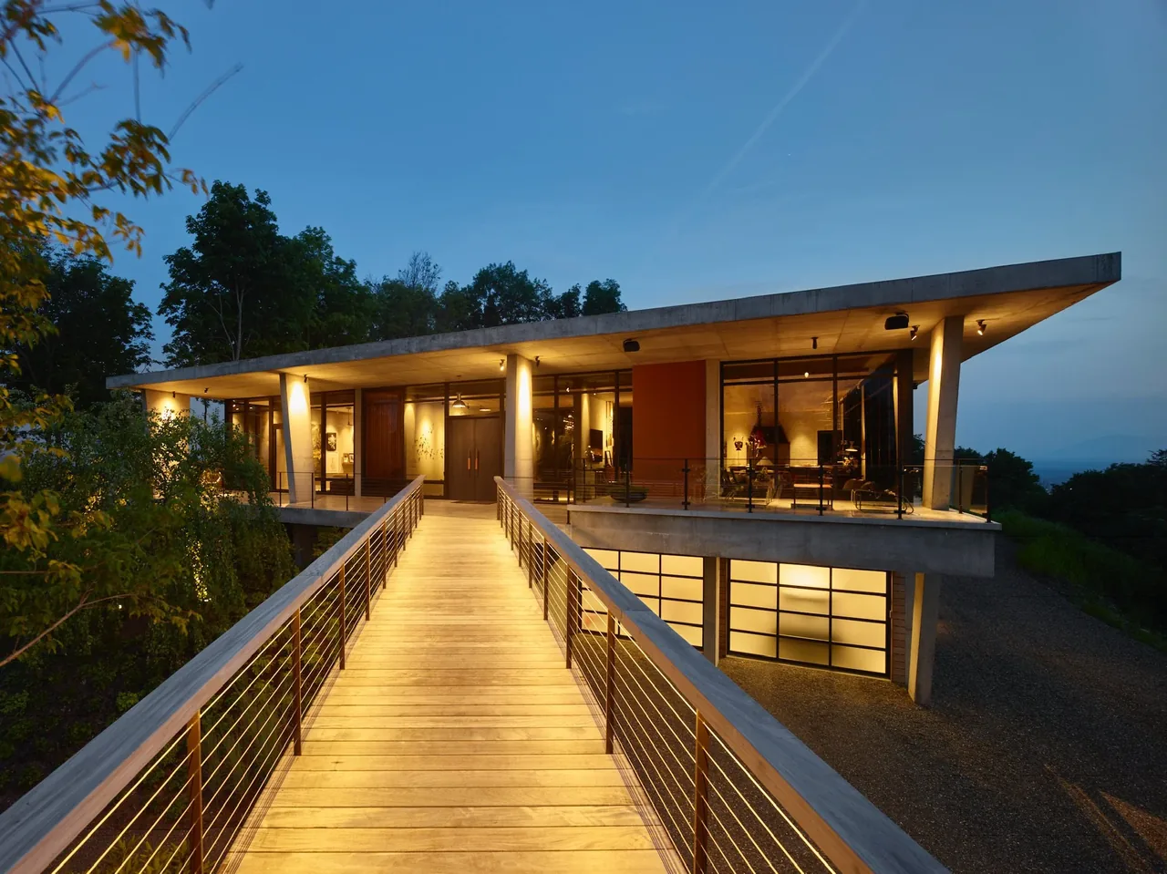 View of a modern concrete house at dusk, with elevated wooden walkway leading to the entrance and warm interior lights.