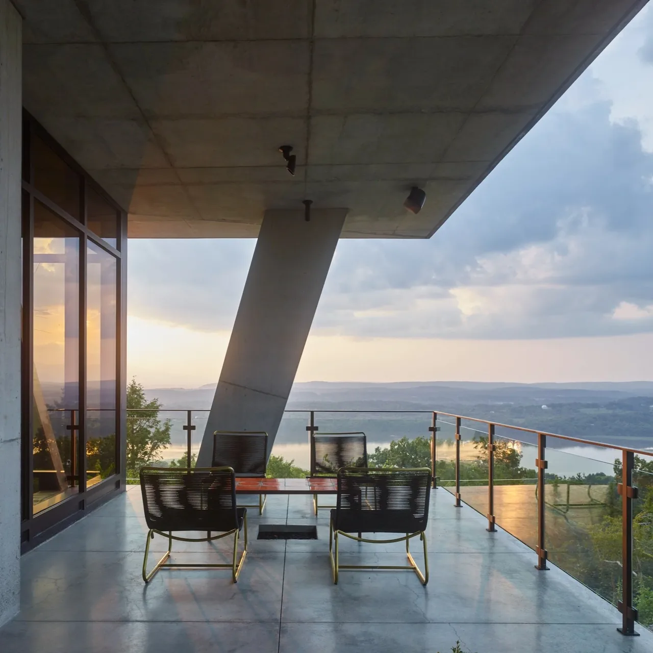 Concrete patio with modern black mesh chairs and glass railing overlooking water and hills at sunset.