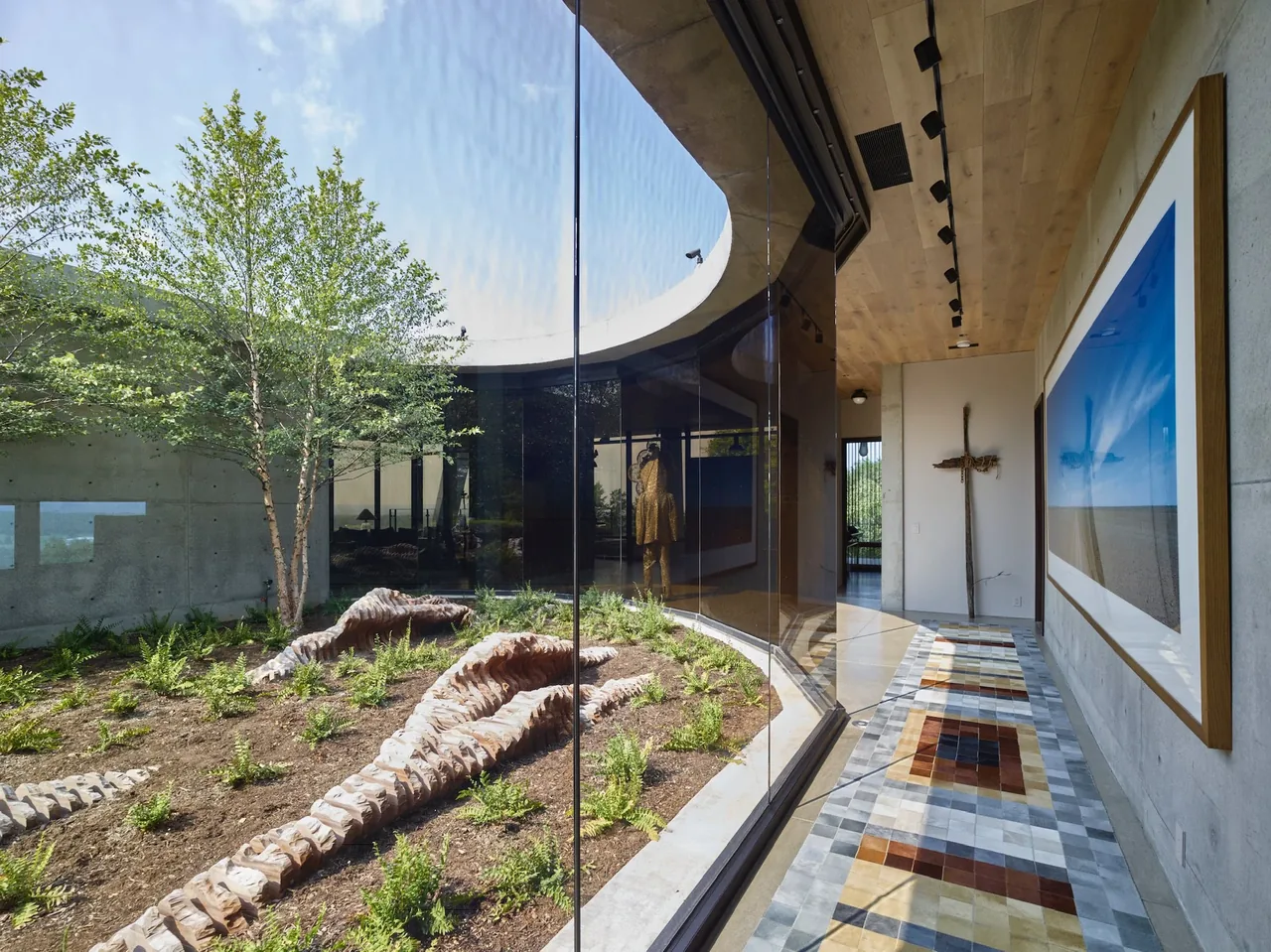 Indoor garden with tree and sculptures viewed through a curved glass wall from a corridor with patterned tile floor and track lighting.