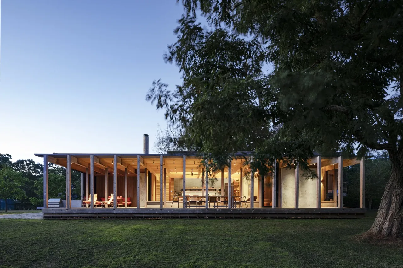 Low modern wood-framed house at dusk with large glass windows and illuminated interior.