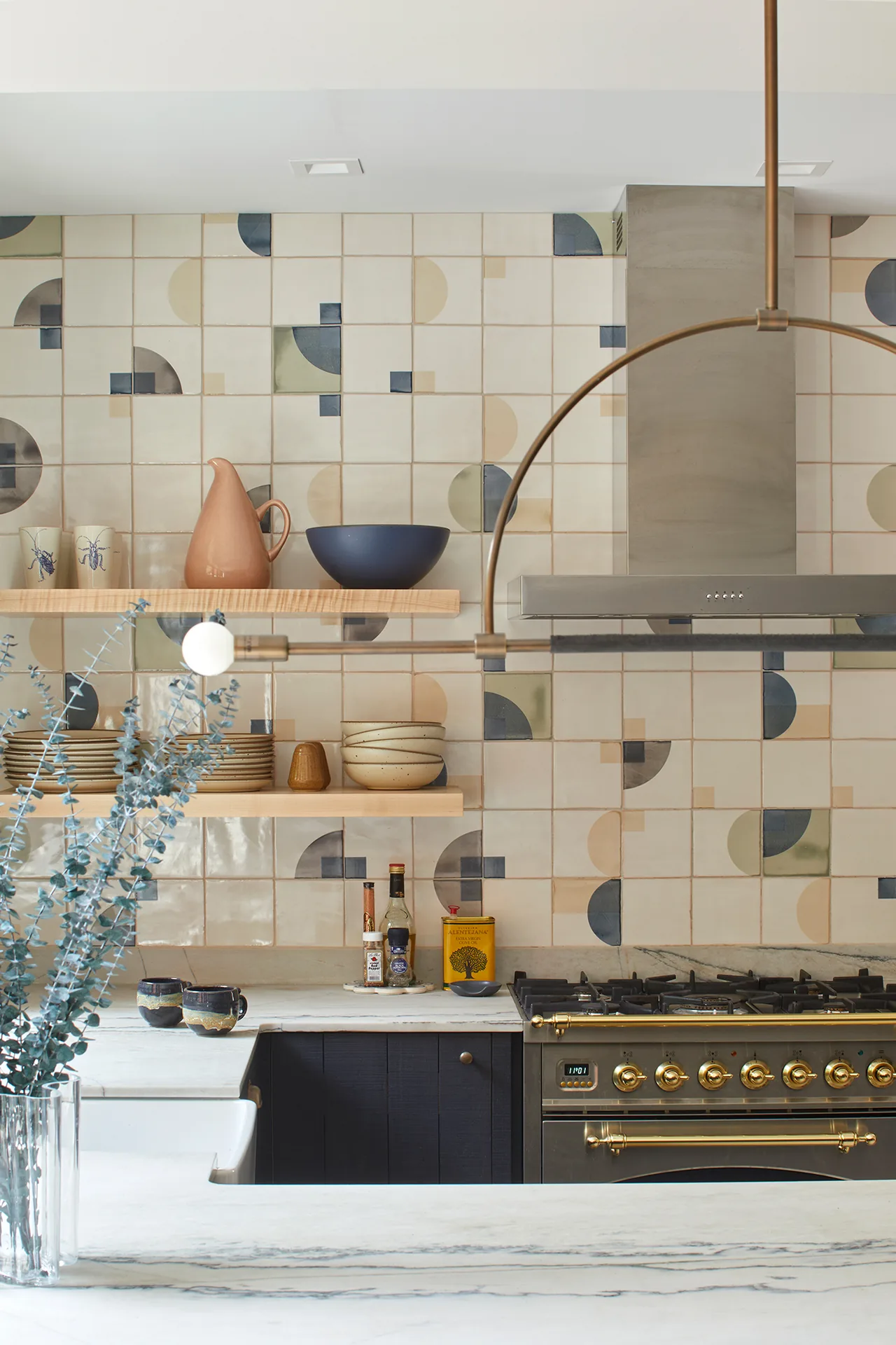 Kitchen with a marble countertop, geometric patterned tile backsplash, and brass-accented stove with stainless steel vent hood.