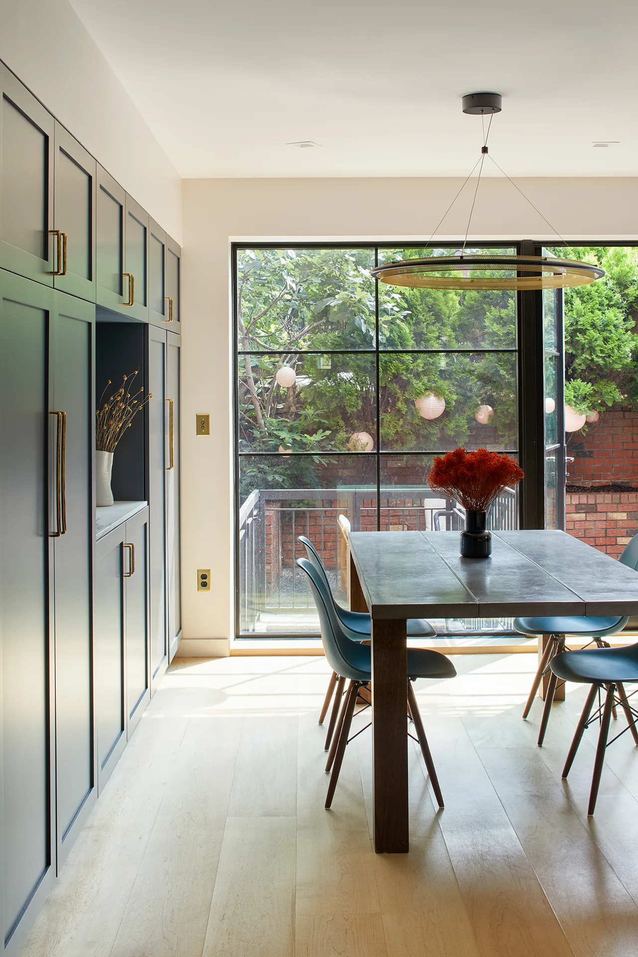 The Greenpoint Residence dining area features blue chairs, a wooden table, and a large window with black frames overlooking an outdoor space.