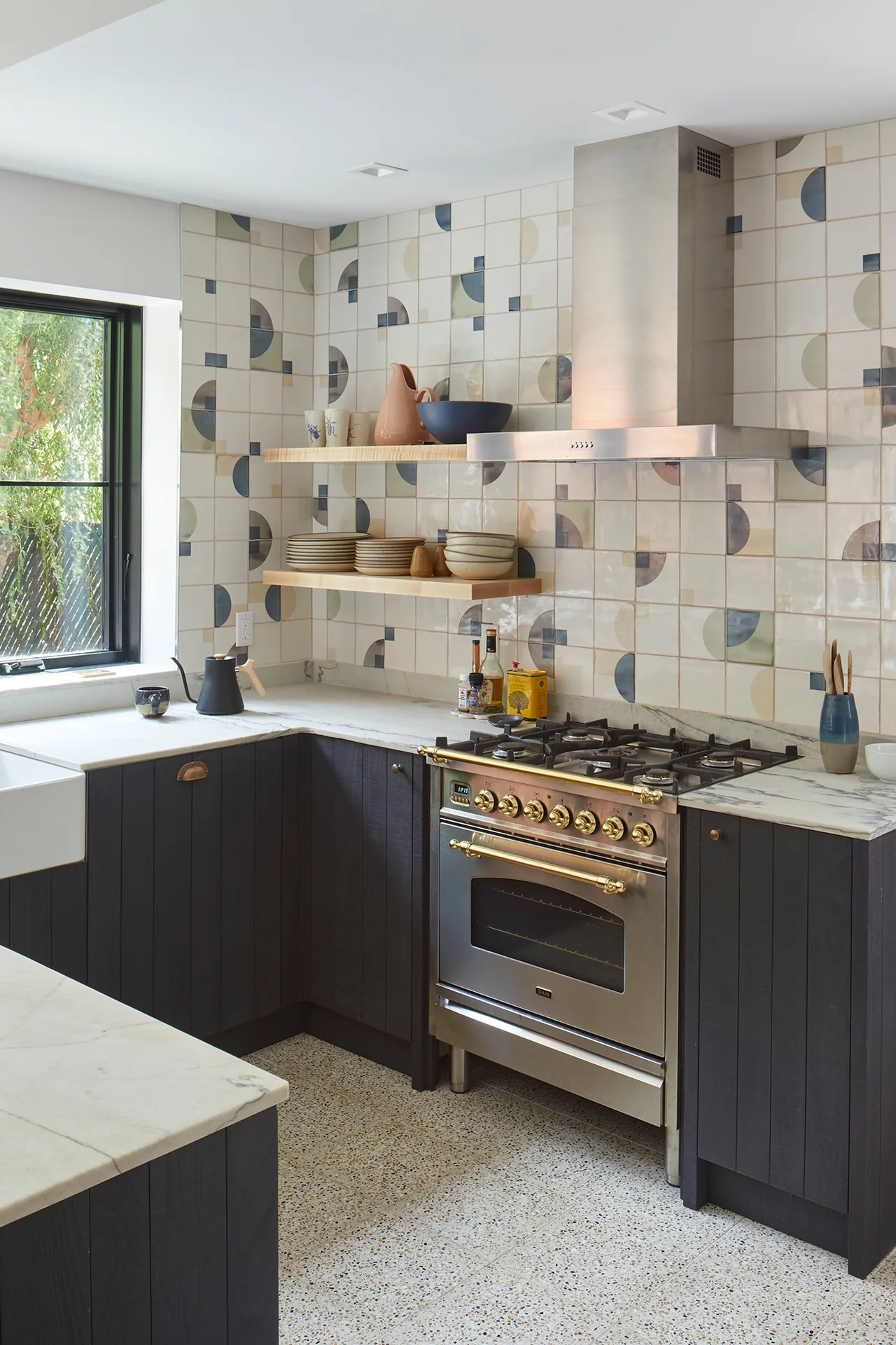 Kitchen area with geometric patterned tile backsplash, open wooden shelves, and a stainless steel gas range with brass handles.