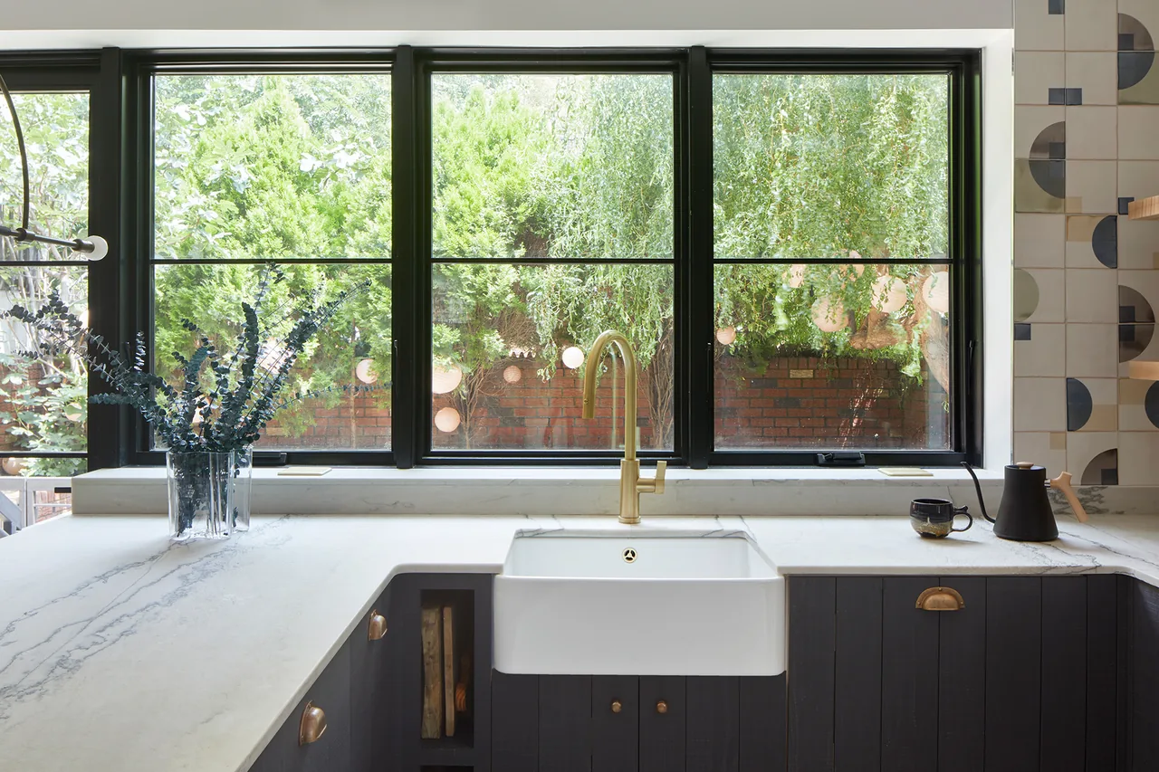 Kitchen area featuring white farmhouse sink with brass faucet below a large black-framed window.