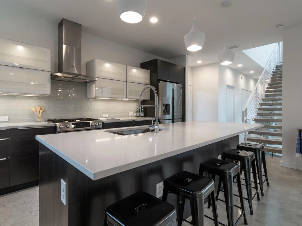 Kitchen with quartz island countertop, black metal stools, stainless steel refrigerator, and a nearby floating staircase.