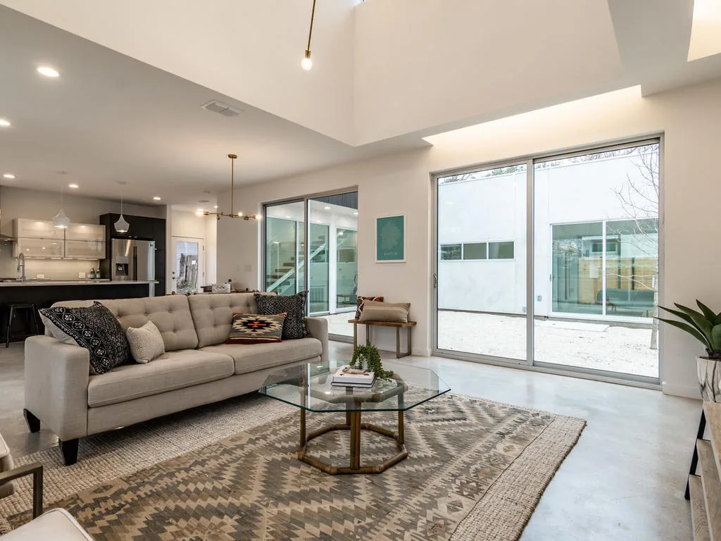 Open living space features a beige sofa, glass coffee table, and sliding glass doors to a white courtyard.