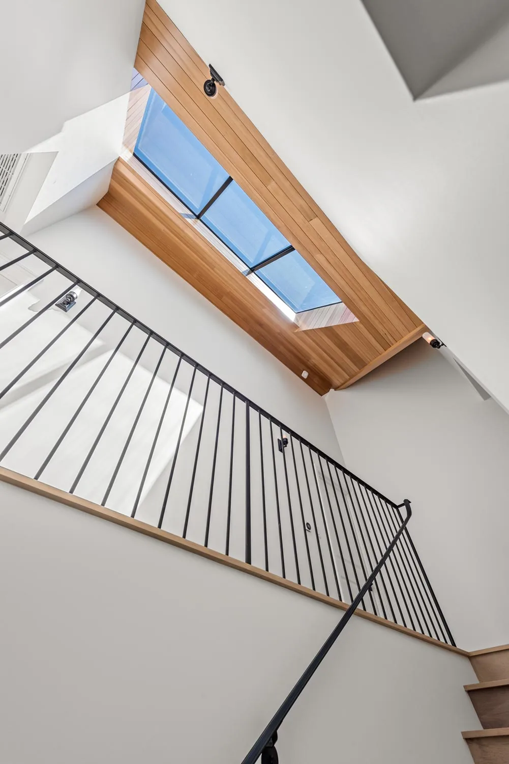 Skylight with wood paneling frames the ceiling above a stairwell with a black metal railing.