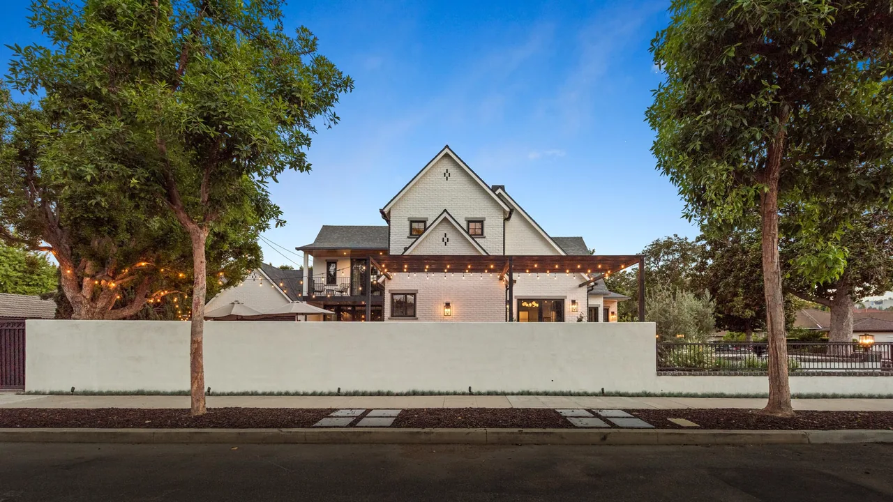 A white brick house with multiple steep gable roofs and a courtyard wall featuring string lights, taken at dusk.