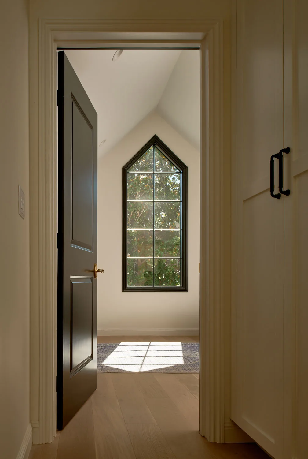 A small room with a vaulted ceiling features a tall pointed window framed in black with natural light casting a shadow on a rug.